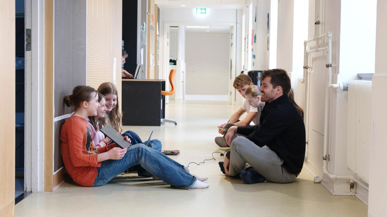 Das Bild zeigt eine Schulumgebung in Dänemark. Im Vordergrund sitzen mehrere Schüler auf dem Boden eines hellen, modernen Flurs. Es sind insgesamt fünf Personen sichtbar: drei Mädchen und zwei Jungen. Die Mädchen sieht man, wie sie auf einem Laptop und einem Tablet arbeiten. Sie haben lässige Kleidung an; eines der Mädchen trägt ein oranges Oberteil mit einem Muster.   Die Jungen sind auf der rechten Seite positioniert. Einer von ihnen hält ein Smartphone, während der andere auf ein Tablet schaut. Die Schüler scheinen in einem entspannten Moment miteinander zu kommunizieren und zu lernen.   Im Hintergrund sind Möbel und Einrichtungen der Schule zu sehen, darunter ein Tisch mit einem Stuhl, ein weiteres Gerät und Wände in hellen Farben. Das Licht ist natürlich und strömt durch Fenster, was dem Raum eine freundliche Atmosphäre verleiht. Die Anordnung der Personen und der Umgebung deutet auf eine kreative Lernsituation hin.