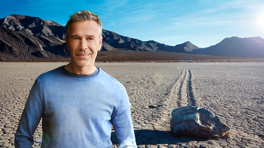 Dirk Steffens kniet auf einem ausgetrockneten See in der Wüste und schaut in die Kamera. Er hält einen Stein mit einer Bohrung in den Händen.