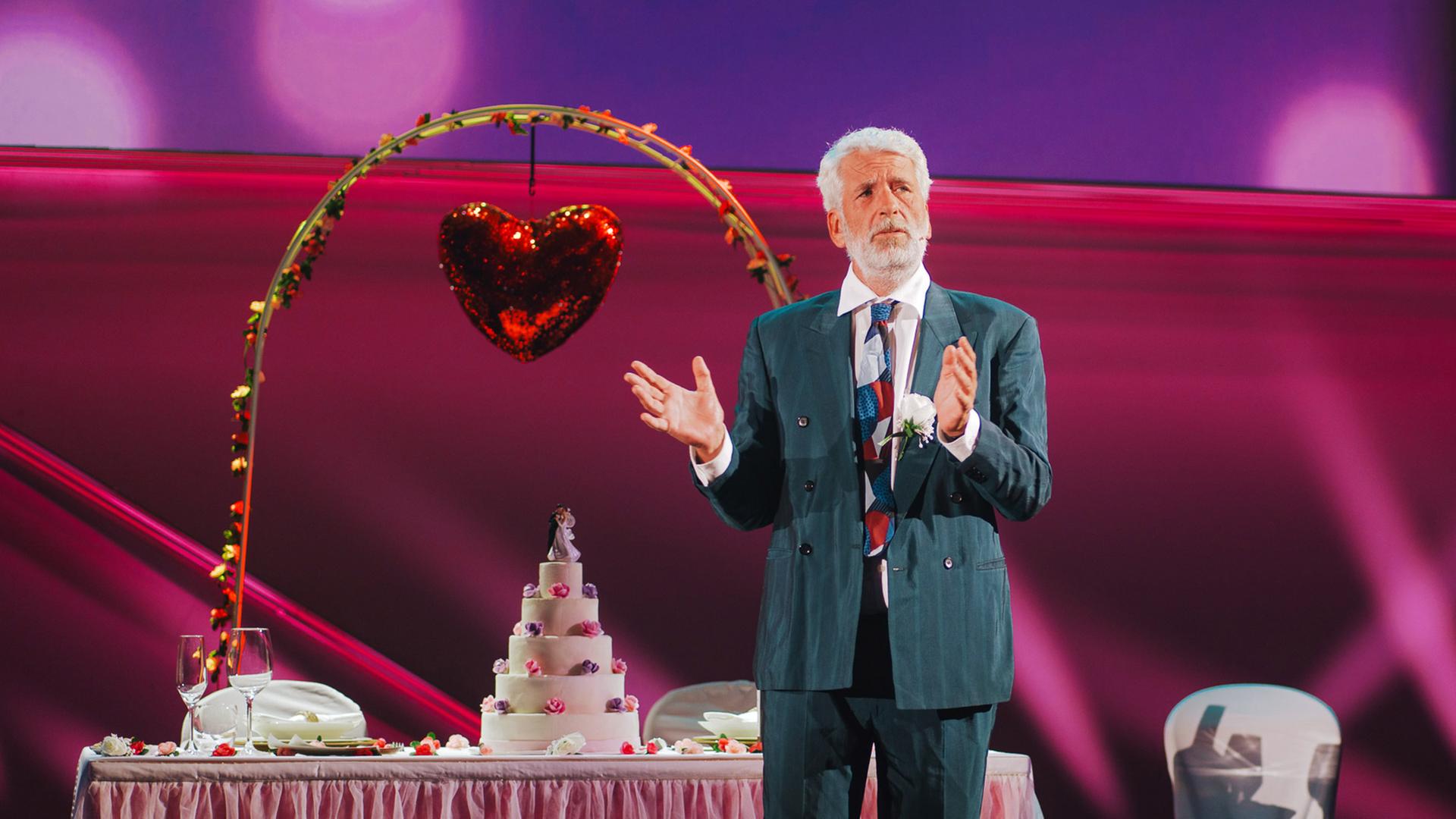 Dirk Stermann steht im Anzug auf der Bühne. Der Hintergrund ist rosa und es ist ein Tisch mit Torte und Gläsern aufgebaut.