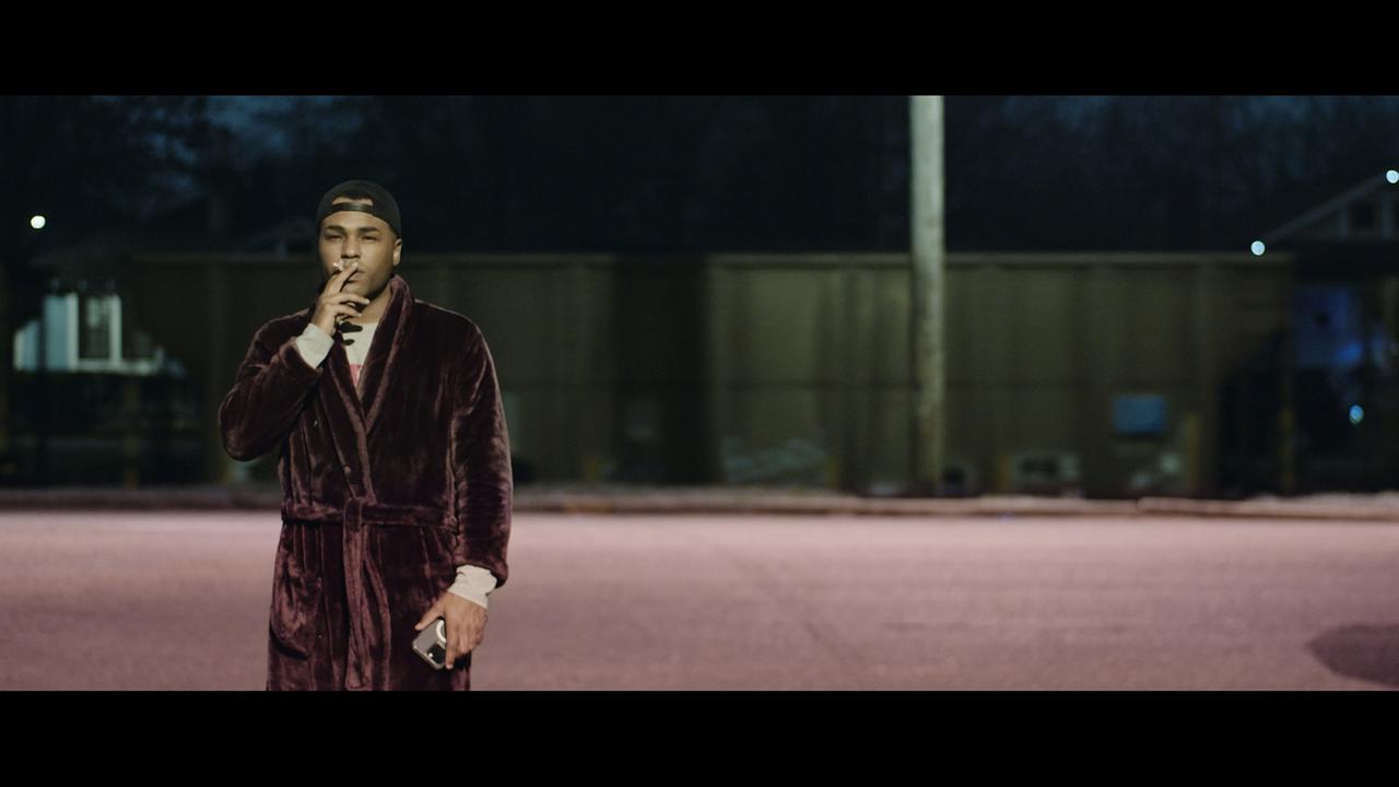 Ein junger Mann in Bademantel steht in der Abenddämmerung vor einer Bahnstrecke und raucht. Hinter ihm fährt ein langer Güterzug entlang.