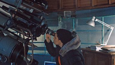 Amena steht am Teleskop einer Sternwarte, hat ein Auge zugekniffen und schaut mit dem anderen in das Okular.