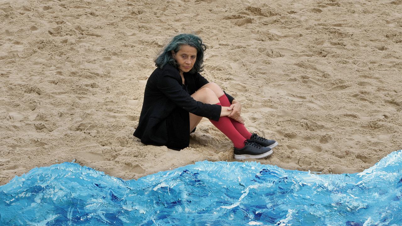 Eine Frau sitzt im Sand. Im unteren Bildrand gemaltes Wasser.