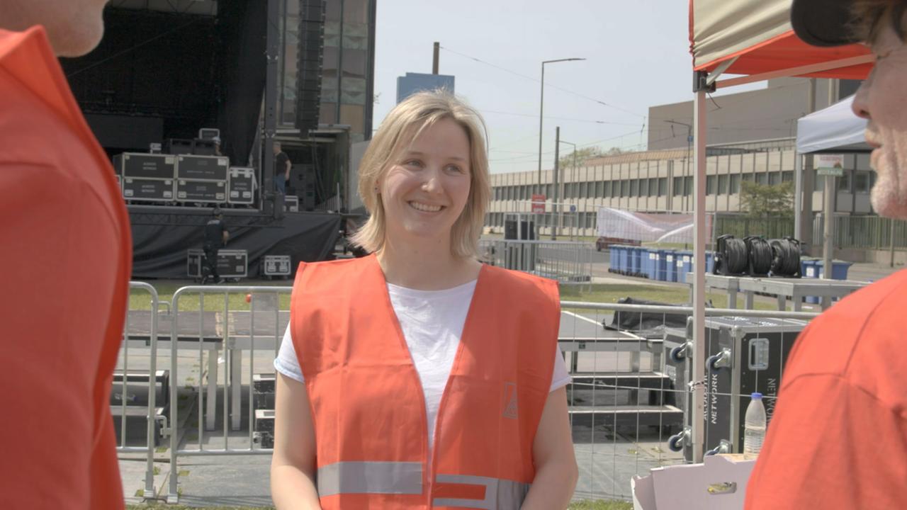 Eine junge Frau mit T-Shirt und Warnweste steht mit zwei Kollegen vor einer großen Bühne zusammen.