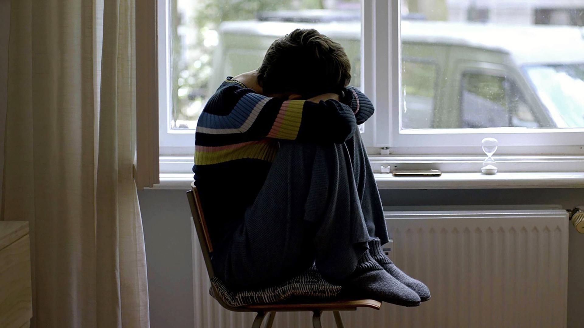 Junge Frau sitzt mit angezogenen Beinen auf einem Stuhl an einem Fenster.