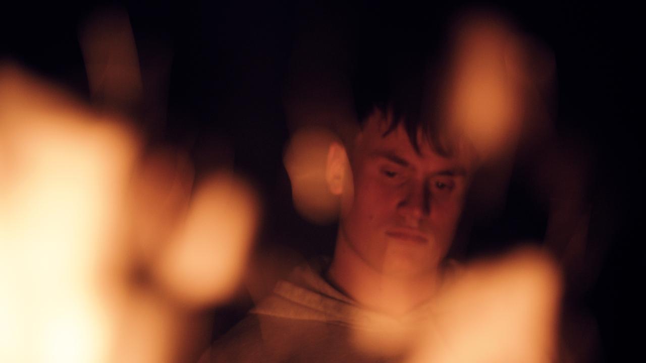 Nahaufnahme eines jungen Mannes, im Vordergrund Flammen eines Lagerfeuers.