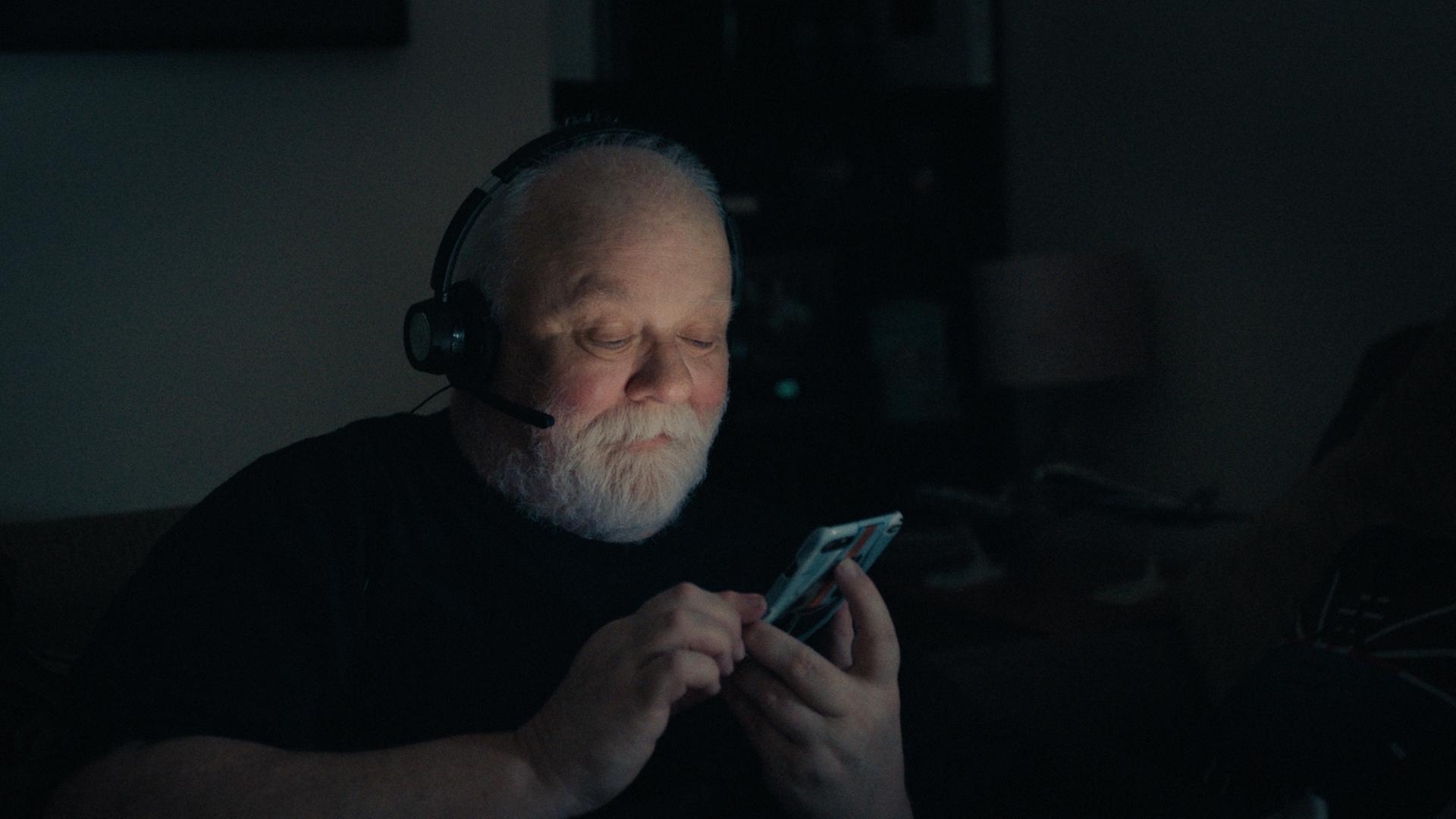 Ein älterer Mann mit glatze und grauem Bart trägt ein Headset und schaut lächelnd auf ein Handydisplay, das ihn im Dunkeln anleuchtet.