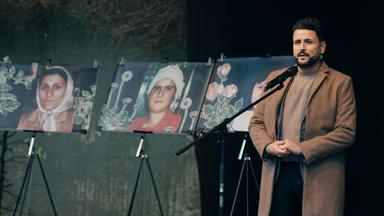 Ein Mann, der einen braunen Mantel trägt, steht rechts von drei Gedächtnisfotografien, die drei Frauen zeigen.