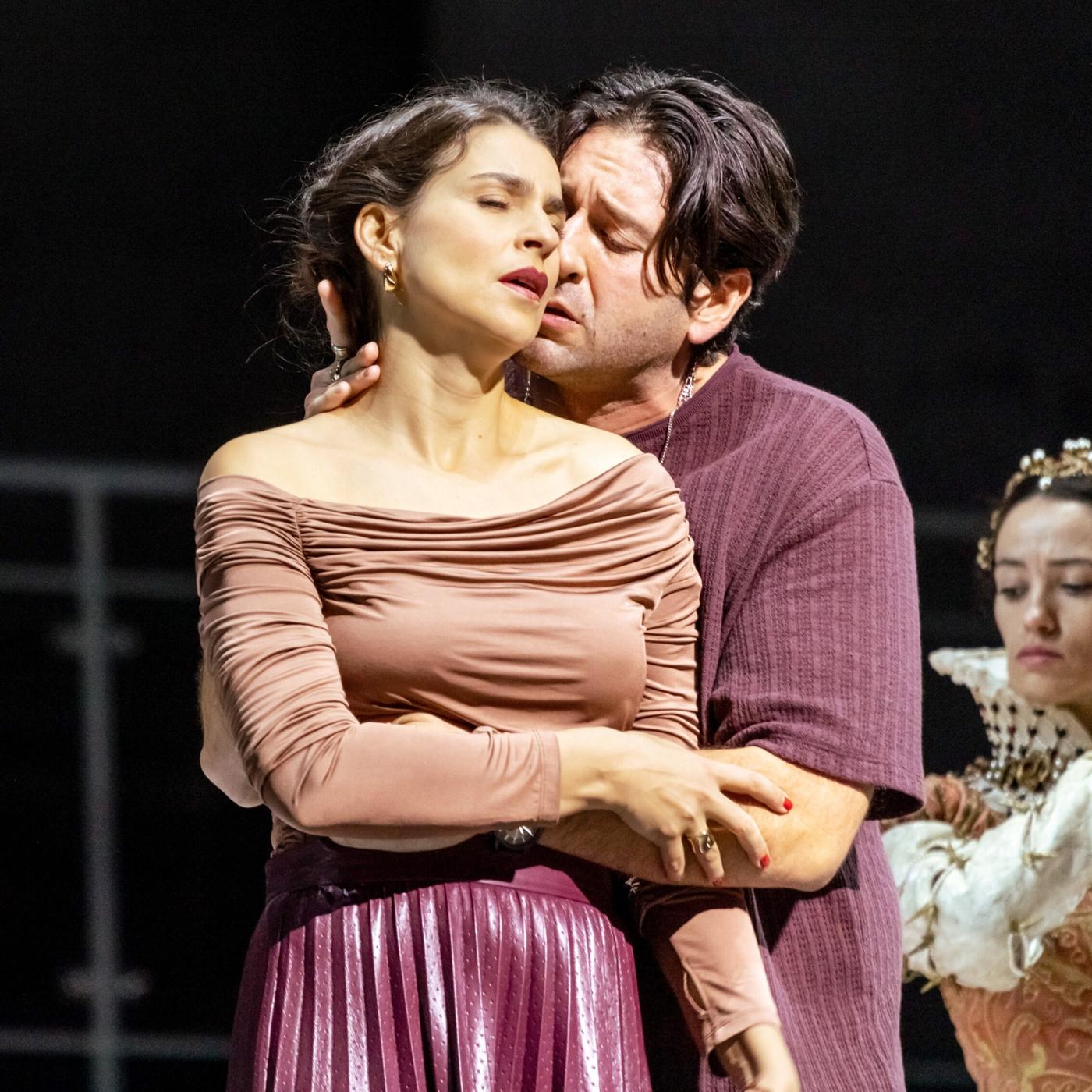 Das Bild zeigt eine Szene aus der Oper „Don Carlo“, die in der Wiener Staatsoper aufgeführt wird. Im Vordergrund sind zwei Hauptfiguren zu sehen. Die Frau trägt ein figurbetontes, rosa Oberteil und einen schimmernden, lila Rock; sie hat leicht geöffneten Mund und wirkt emotional. Der Mann, der sie umarmt, trägt ein lockeres, weinrotes Oberteil und hat einen konzentrierten Gesichtsausdruck, während er ihr nahekommt. Im Hintergrund befindet sich eine weitere Figur in einer aufwendig gestalteten Kostümierung, die einen historischen Stil aufweist. Sie steht etwas abseits und wirkt in ihrer Pose distanziert. Die Bühne wirkt spartanisch, mit einem dunklen Hintergrund, was den Fokus auf die Schauspieler lenkt. Das Bild vermittelt intensive Emotionen und die Dramatik der opernhaften Darbietung.