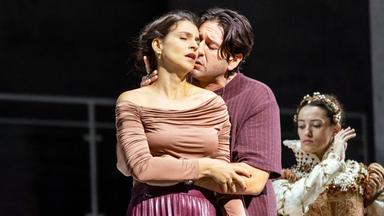 Das Bild zeigt eine Szene aus der Oper „Don Carlo“, die in der Wiener Staatsoper aufgeführt wird. Im Vordergrund sind zwei Hauptfiguren zu sehen. Die Frau trägt ein figurbetontes, rosa Oberteil und einen schimmernden, lila Rock; sie hat leicht geöffneten Mund und wirkt emotional. Der Mann, der sie umarmt, trägt ein lockeres, weinrotes Oberteil und hat einen konzentrierten Gesichtsausdruck, während er ihr nahekommt. Im Hintergrund befindet sich eine weitere Figur in einer aufwendig gestalteten Kostümierung, die einen historischen Stil aufweist. Sie steht etwas abseits und wirkt in ihrer Pose distanziert. Die Bühne wirkt spartanisch, mit einem dunklen Hintergrund, was den Fokus auf die Schauspieler lenkt. Das Bild vermittelt intensive Emotionen und die Dramatik der opernhaften Darbietung.