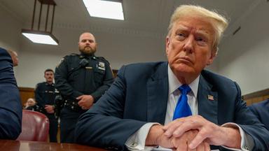 Ein älterer Mann mit gelben Haaren und erstem Gesicht, gekleidet in einen dunkelblauen Anzug mit blauer Krawatte, sitzt an einem Tisch, hinter ihm steht ein Beamter in Uniform.