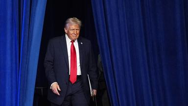 Ein älterer Mann im blauen Anzug mit roter Krawatte tritt durch einen geöffneten blauen Vorhang.