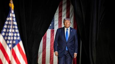 Ein älterer Mann im blauen Anzug tritt durch einen schwarzen Vorhand. Im Hintergrund sind Flaggen der USA zu sehen.