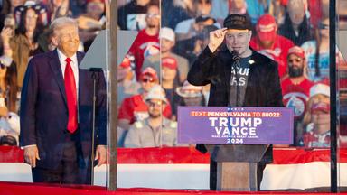 Ein älterer Mann in blauem Anzug mit roter Krawatte und ein Mann mit Baseballkappe und legerer Kleidung stehen hinter Panzerglas an Rednerpulten. Hinter ihnen ist Publikum zu sehen.