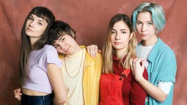 Das Bild zeigt vier Frauen, die in einer Gruppenaufnahme stehen. Sie sind vor einem einfarbigen, leicht strukturierten Hintergrund in einem sanften Rotton positioniert. Jede Frau trägt einen unterschiedlichen Kleidungstil und ist in verschiedenen Farben gekleidet:  1. Die erste Frau links hat lange, dunkle Haare und trägt ein lila kurzes Oberteil mit einer dunkelblauen Hose. 2. Die zweite Frau steht etwas vor der ersten und hat kurze, dunkle Haare. Sie trägt ein gelbes Oberteil über einem weißen T-Shirt und eine orangefarbene Hose. 3. Die dritte Frau hat langes, hellbraunes Haar und trägt einen roten Latzhose über einem roten Oberteil. 4. Die vierte Frau im Bild hat kurze, hellblaue Haare und trägt ein türkisfarbenes Oberteil über einem weißen T-Shirt.  Die Frauen haben verschiedene Posen eingenommen und lächeln leicht oder schauen in die Kamera. Ihre Körperhaltung und der freundliche Gesichtsausdruck vermitteln eine entspannte und positive Stimmung.
