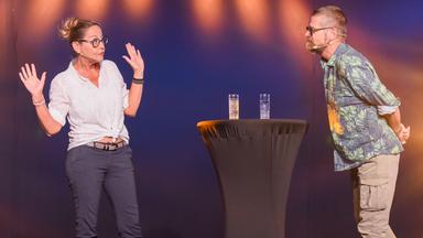 Das Bild zeigt zwei Personen auf einer Bühne. Zu sehen sind Monica Weinzettl und Gerold Rudle, die in einer humorvollen Interaktion miteinander stehen. Monica Weinzettl trägt ein helles, kurzärmliges Hemd und dunkle, bequeme Hose. Ihr Haar ist zu einem lockeren Dutt frisiert, und sie hat eine Brille auf. Gerold Rudle steht etwas seitlich mit dem Rücken zur Kamera und trägt ein farbenfrohes Hemd mit einem Pflanzenmuster und helle, lockere Hosen.   Zwischen den beiden steht ein runder Tisch mit zwei Gläsern Wasser darauf. Die Bühne ist in schwaches Licht getaucht, mit farbigen Lichtern, die eine warme Atmosphäre schaffen, und der Hintergrund ist dunkel.  Die Personen scheinen in eine lebhafte Rede oder Vorstellung vertieft zu sein, wobei Weinzettl ihre Arme ausbreitet, als ob sie einen Punkt betonen möchte. Die Szene vermittelt eine entspannte und humorvolle Stimmung.