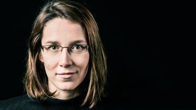 Eine lächelnde Frau mit langen Haaren und Brille schaut den Betrachter direkt an.