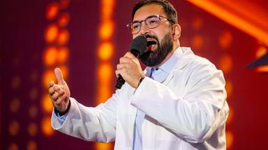 Dr. Emir trägt einen weißen Kittel und ein blaues Hemd darunter. In seiner linken Hand hält er das Mikrofon. Der Hintergrund ist rot beleuchtet. Dr. Emir trägt eine Brille.