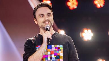 Dr. Pop steht in der Mitte des Bildes. Er trägt einen schwarzen Pullover mit bunten Vierecken darauf. In seiner rechten Hand hält er ein Mikrofon. Seine linke Hand hat er in die Hosentasche gesteckt. Im Hintergrund sind Scheinwerfer zu sehen.