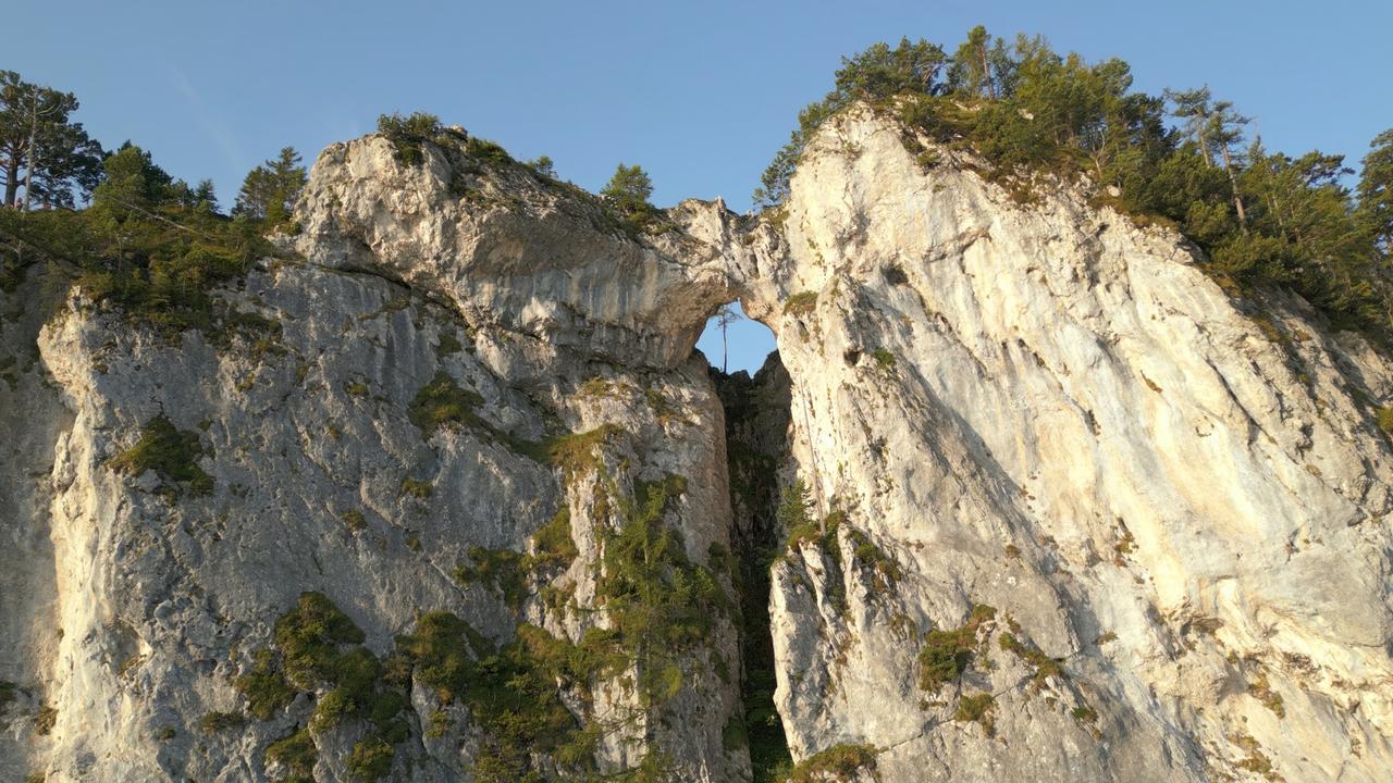 Das Bild zeigt die Drachenwand, einen markanten Berg im Mondseeland in Österreich. Besonders auffällig ist das große Loch, das sich in der vertikalen Felswand befindet. Die Felsoberfläche ist rau und unregelmäßig, mit verschiedenen Tönen von Grau und Beige. In einigen Bereichen wachsen kleine grüne Bäume und Sträucher, die sich an die Felsen schmiegen.   Der Himmel ist klar und blau, was dem Bild eine helle und frische Atmosphäre verleiht. Diese Landschaft wird häufig von Kletterern besucht, die den anspruchsvollen Klettersteig nutzen. Die Drachenwand ist von zahlreichen Mythen umgeben, was in der Region bekannt ist.