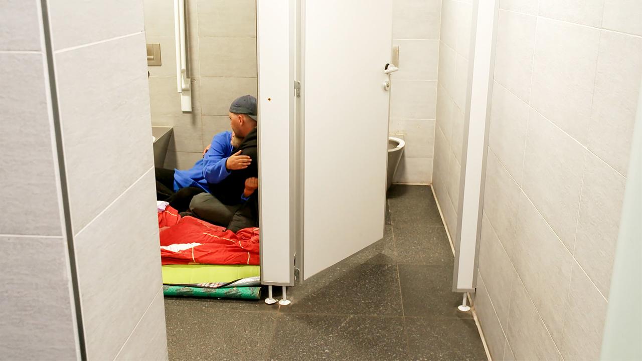 Das Bild zeigt den Innenbereich eines modernen, öffentlichen Toilettenraums. Auf der linken Seite ist eine graue Wand mit einer Trennwand zu sehen, die einen kleinen Bereich abgrenzt. Auf dem Boden liegt eine orangefarbene und eine grüne Decke, auf denen zwei Personen sitzen.   Eine Person, die in einem blauen Oberteil gekleidet ist, umarmt eine andere Person, die mit einem neutralen Kleidungsstil bekleidet ist. Im Hintergrund ist eine Toilette sichtbar, die an die Wand angestrichen ist. Die Wände sind mit hellen Fliesen ausgekleidet, und der Boden ist mit dunklem Material gestaltet. Das Licht im Raum ist hell und sorgt für eine klare Sicht. Es gibt keine Fenster, was den Raum etwas bedrückend erscheinen lässt.  Das Bild vermittelt die Atmosphäre eines temporären Schlafplatzes, in dem sich die beiden Personen aufhalten.