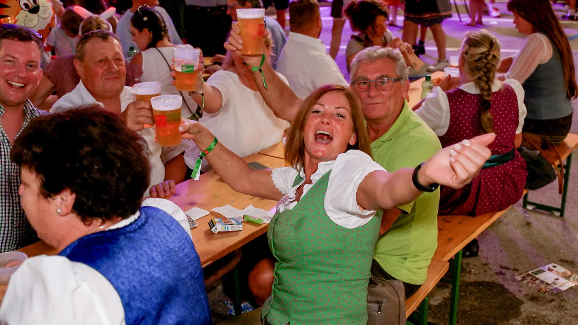 Das Bild zeigt eine lebhafte Szene auf einem Fest in Windischgarsten. Mehrere Personen sitzen an langen Holztischen und halten Becher mit Bier in den Händen. In der Mitte des Bildes ist eine Frau mit braunen Haaren, die in einem traditionellen Dirndlkleid (grünes Mieder und weiße Bluse) gekleidet ist. Sie lächelt breit und hat ihren Arm nach oben gestreckt, als ob sie feiern würde.   Um sie herum sind weitere Menschen sichtbar, einige ebenfalls in Trachten bekleidet. Der Hintergrund ist unklar, aber es gibt eine Atmosphäre von Geselligkeit und Feierlichkeit. Einige Personen heben ihre Becher, während andere in Gespräche vertieft sind. Der Boden scheint ein festlicher Ort zu sein, möglicherweise mit bunten Lichtern und Dekorationen, passend zu einem traditionellen Fest.   Es ist eine fröhliche Umgebung, in der die Leute Spaß haben und die Atmosphäre ausgelassen wirkt.