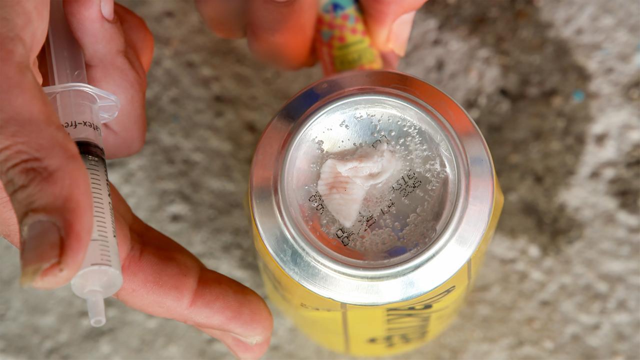 Das Bild zeigt eine Hand, die eine Spritze hält, während die andere Hand eine Dose mit einem silbernen Deckel hält, auf dem sich eine körnige Substanz befindet. Die Dose hat eine gelbe Farbe mit schwarzer Schrift. Die Substanz auf dem Deckel sieht kristallin aus und könnte eine Drogenmarke sein. Der Hintergrund des Bildes ist unscharf und besteht aus grauem Beton oder einem ähnlichen Material. Lichtreflexionen sind auf der Dose sichtbar, die die glatte Oberfläche hervorheben. Es handelt sich um eine bildliche Darstellung des Drogenkonsums.