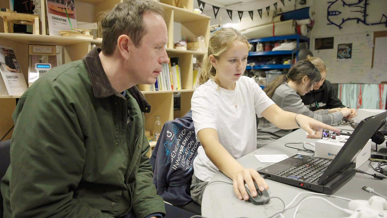 Das Bild zeigt eine Szene in einem Raum, in dem junge Menschen, insbesondere Mädchen, lernen, wie man mit Drohnen arbeitet. Im Vordergrund sind zwei Personen zu sehen: ein Mann und ein Mädchen. Der Mann sitzt links und trägt eine grüne Jacke. Er schaut konzentriert auf den Laptop und scheint mit dem Mädchen zu sprechen. Das Mädchen sitzt rechts neben ihm. Sie hat lange, lockige Haare und trägt ein weißes T-Shirt. Sie ist dabei, auf einen Punkt auf dem Laptop hinzuweisen, während sie eine Hand mit einem kleinen Gerät hält.  Im Hintergrund sind weitere Jugendliche sichtbar, die an ähnlichen Projekten arbeiten. Der Raum ist mit Regalen und verschiedenen Materialien ausgestattet, die auf kreative Aktivitäten hinweisen. An der Wand sind einige Zeichnungen oder Plakate sichtbar. Es herrscht eine konzentrierte und kreative Atmosphäre, die die Begeisterung der jungen Menschen für Technik und Drohnen zeigt.