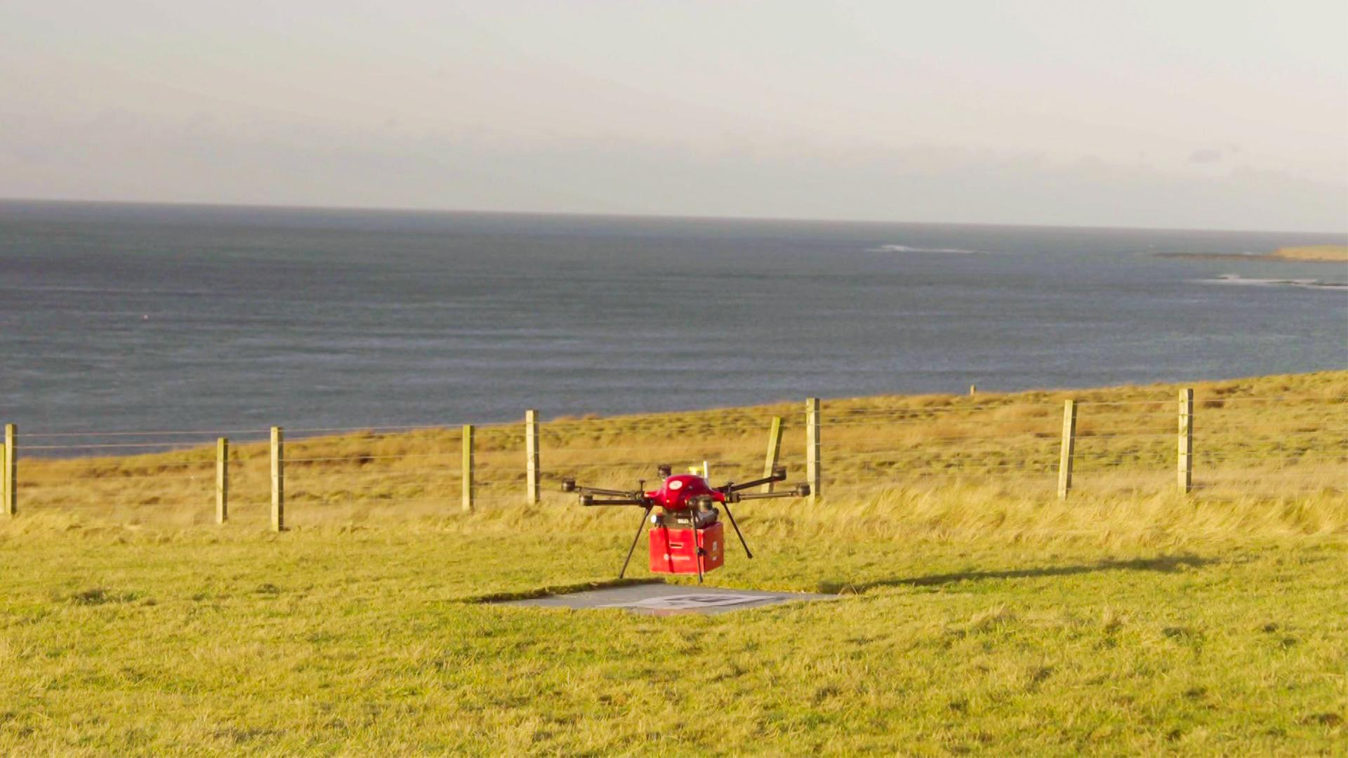 Das Bild zeigt eine leichte, quadratische Drohne, die in einem offenen, grünen Feld auf den schottischen Orkney-Inseln positioniert ist. Die Drohne hat ein rotes Gehäuse und ist über einer markierten Landezone platziert. Im Hintergrund erstreckt sich das Meer, das mit einem sanften Wellengang zu sehen ist. Die Küstenlinie ist am Horizont sichtbar, während sich eine Reihe von Holzpfählen entlang des Feldes zieht, die eine Umzäunung bilden. Der Himmel ist größtenteils klar, mit einigen Wolken, und es scheint Tageslicht zu sein.   Die Szene vermittelt ein Gefühl von Abgeschiedenheit und der technischen Anwendung von Drohnen in ländlichen Gebieten, wo sie zum Transport von wichtigen Gütern eingesetzt werden.