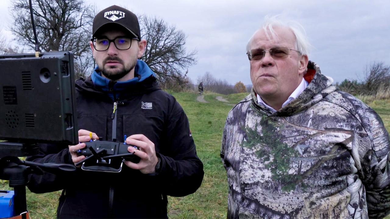 Das Bild zeigt zwei Männer, die an einem offenen, grünbewachsenen Feld stehen. Der Himmel ist bewölkt und es herrscht eine graue Atmosphäre. Der jüngere Mann trägt eine schwarze Jacke und eine schwarze Baseballmütze mit dem Aufdruck "DYNAT". Er hält einen Controller in der Hand und schaut konzentriert auf einen Bildschirm, der vor ihm auf einem Stativ steht. An dem Bildschirm sind einige Antennen angebracht.  Der ältere Mann steht direkt neben ihm und trägt einen camouflagefarbenen Hoodie. Er hat graue Haare, trägt eine Brille und sieht ernst aus, während er in die gleiche Richtung wie der jüngere Mann blickt. Im Hintergrund ist ein weiterer Mensch auf einem Weg zu erkennen, der durch das Feld führt. Die Umgebung wirkt ländlich und unberührt, mit wenigen Bäumen und Sträuchern.