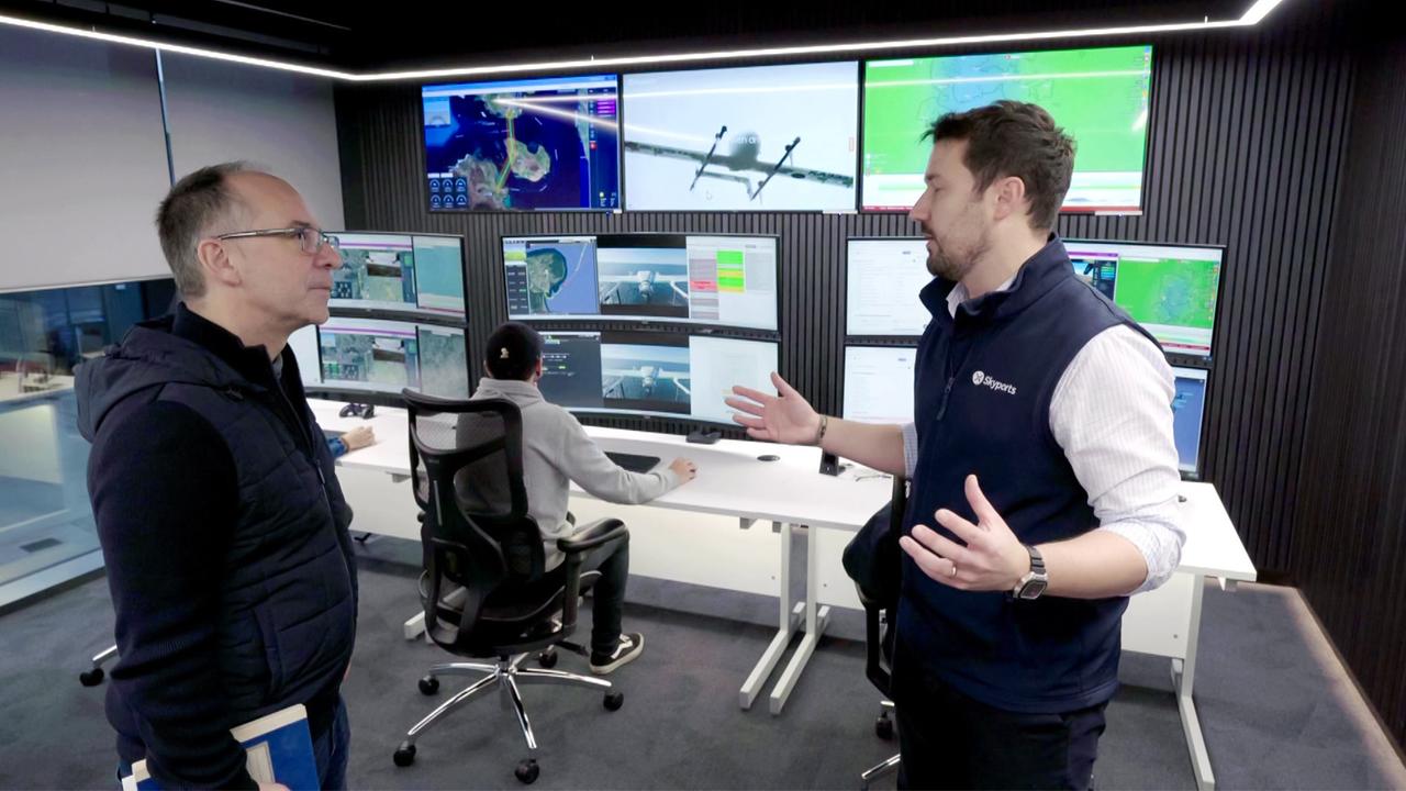 Das Bild zeigt eine moderne Kontrollzentrale, in der Drohnen überwacht und gesteuert werden. Am unteren Rand des Bildes stehen zwei Männer, die sich unterhalten. Der Mann links trägt eine dunkle Weste und hält etwas in der Hand. Der Mann rechts trägt eine dunkle Weste mit einem hellen Hemd darunter.   Im Hintergrund sitzen zwei Personen an Tischen mit Monitoren. Ein Monitor zeigt ein Luftbild mit einer Flugroute, während andere Bildschirme Daten und Karten darstellen. Die Wände des Raumes sind mit großen Bildschirmen ausgestattet, auf denen verschiedene Informationen über die Drohnen angezeigt werden.  Die gesamte Umgebung wirkt aufgeräumt und modern, mit einer technischen Atmosphäre, die sich auf die Drohnentechnologie konzentriert.