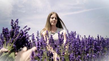 Das Bild zeigt eine junge Frau, die in einem Lavendelfeld steht. Sie hat lange, braune Haare und trägt ein schlichtes, graues T-Shirt. Ihr Gesicht zeigt einen nachdenklichen Ausdruck, während sie von mehreren Lavendelpflanzen umgeben ist, deren violette Blüten lebhaft und prachtvoll in den Vordergrund des Bildes ragen.   Im Hintergrund ist der Himmel teilweise bewölkt, was eine sanfte Beleuchtung erzeugt. Es ist ein harmonisches und frisches Ambiente, das die natürliche Schönheit der Umgebung unterstreicht. Die Szene vermittelt ein Gefühl von Ruhe und der Arbeit in der Natur, insbesondere in Bezug auf die Ernte von Lavendel, der oft mit der Herstellung von Kosmetikprodukten assoziiert wird.
