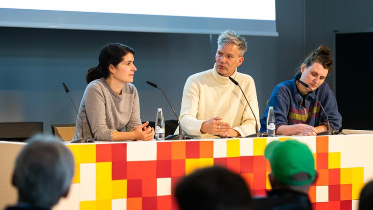Hannah Pilarczyk, Andreas Bolm und Yana Höhnerbach sitzen auf dem 3sat-Extra-Podium der Duisburger Filmwoche 2022.