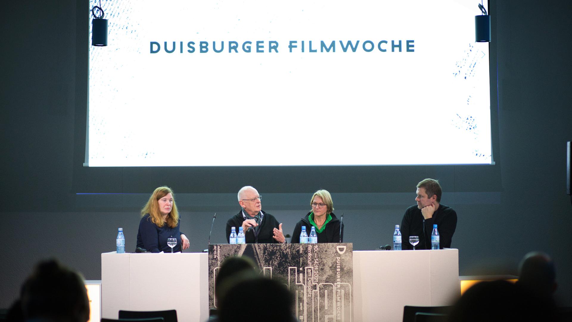 Britt Beyer, Michael Loeken, Ulrike Franke und Matthias Dell sitzen nebeneinander hinter einem Tisch auf einem Podium. Auf einer Leinwand hinter ihnen ist der Schriftzug "Duisburger Filmwoche" projiziert.