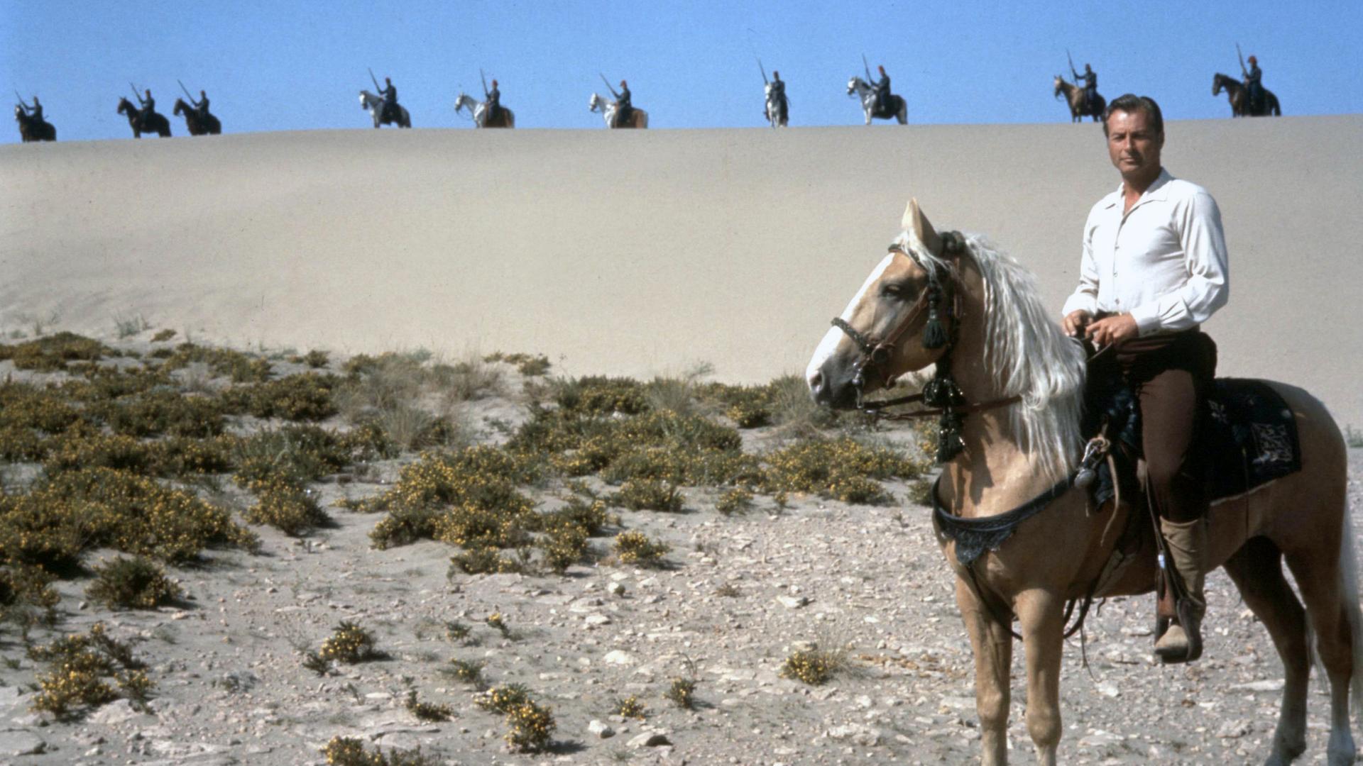 Kara Ben Nemsi (Lex Barker) sitzt auf einem Pferd und schaut in die Ferne. Hinter ihm auf einer Sanddüne sieht man mehrere bewaffnete Krieger, ebenfalls auf Pferden.