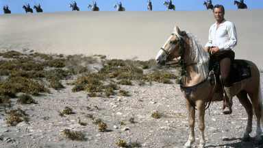 Kara Ben Nemsi (Lex Barker) sitzt auf einem Pferd und schaut in die Ferne. Hinter ihm auf einer Sanddüne sieht man mehrere bewaffnete Krieger, ebenfalls auf Pferden.