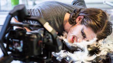 Tänzer Andrew Cummings legt sein Gesicht in eine Metallschüssel mit Daunenfedern. Die Hälfte seines weiß geschminkten Gesichtes ist noch zu sehen.