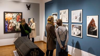 In einem Ausstellungsraum betrachten vier Frauen in Zweiergrüppchen Bilder an den Wänden. In der Mitte des Raums steht ein Holzsarg.