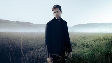 Ein Mann steht auf einer Wiese vor einem verschwommenen Hintergrund von Bergen.