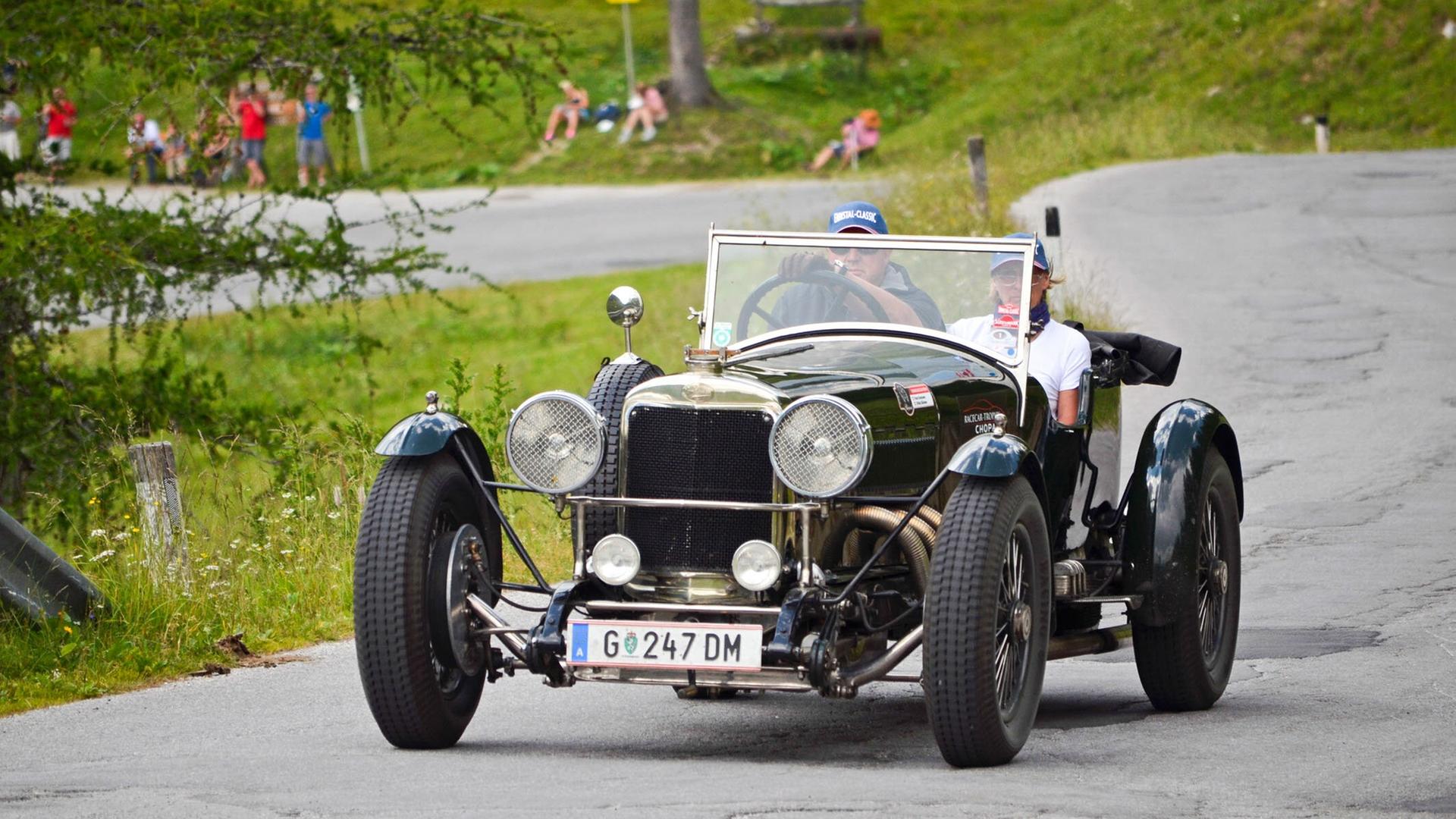 Ein dunkelgrüner Sunbeam Supersport Oldtimer mit zwei Insassen fährt eine Bergstraße entlang, im Hintergrund Zuschauer auf einer Wiese.