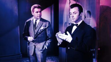 Ein Mann im Anzug hält ein hölzernes Objekt und schaut einen Butler in Livree an, der sich weiße Handschuhe anzieht.