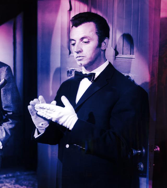 Ein Mann im Anzug hält ein hölzernes Objekt und schaut einen Butler in Livree an, der sich weiße Handschuhe anzieht.