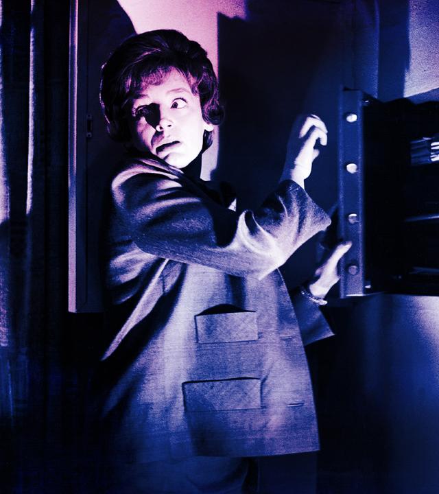 Eine Frau steht vor einem halb geöffneten Wandsafe im Halbschatten, schlaglichtartig wird ihr Gesicht von einer einzelnen Lichtquelle erhellt. Vorsichtig blickt sie über ihre Schulter.