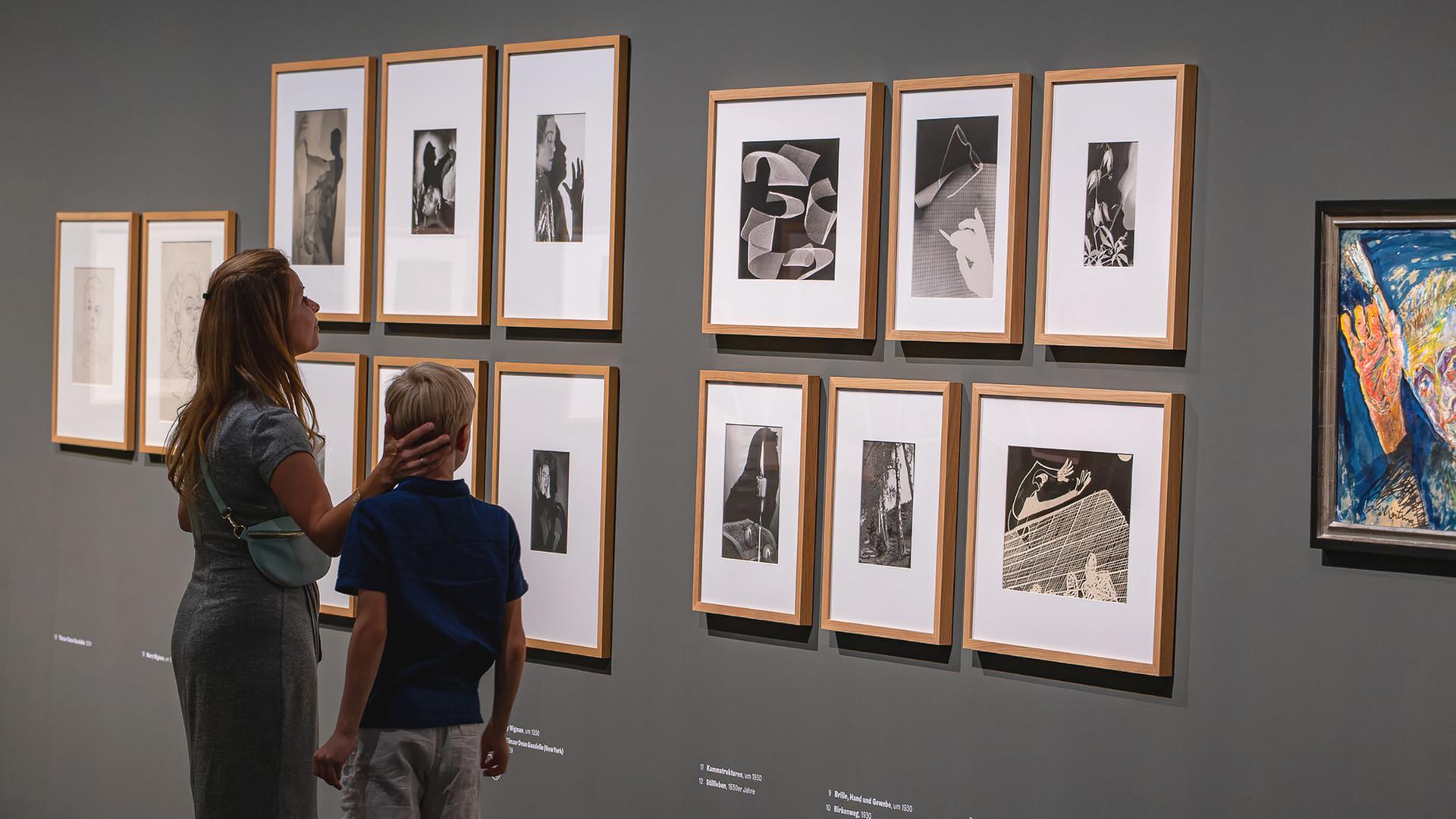 Eine Frau und ein Kind schauen sich Schwarz-Weiß-Fotos und Fotogramme an, die in einem Ausstellungsraum an der Wand hängen.