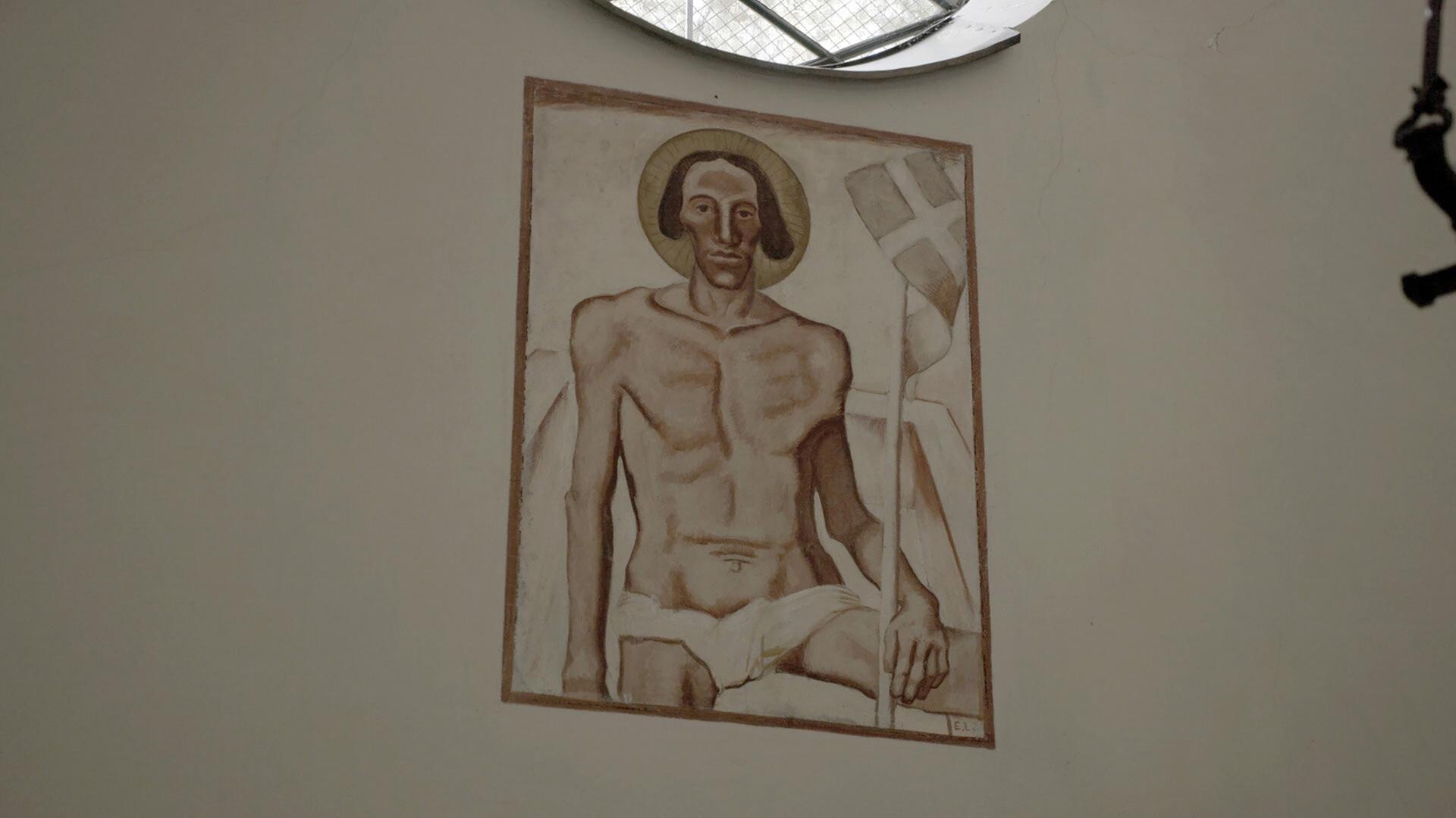 Das Bild zeigt ein Wandgemälde mit einer Darstellung von Christus als Halbfigur. Er sitzt auf einem Stein, hat einen Heiligenschein und hält eine Fahne. Die Farben sind gedämpft und erdtonartig.