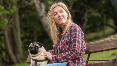 „Ein Mops zum Verlieben“: Sarah (Beattie Edmondson) sitzt mit Mops Patrick auf einer Parkbank. Beide schauen interessiert in die gleiche Richtung.