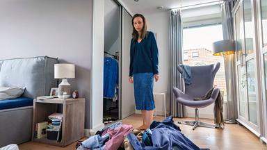 Eine Frau steht vor einem Klamottenhaufen auf dem Boden eines Schlafzimmers.