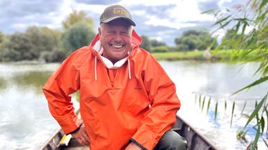 Ein Mann in orangefarbener Regenjacke kniet strahlend in einem kleinen Boot auf einem See