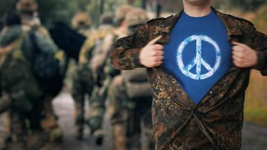 Ein Mann in Flecktarn reißt seine Jacke auf und trägt darunter ein T-Shirt mit einem Peace-Zeichen.