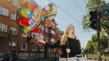 Eine Frau hält mehrere bunte Luftballons mit der Aufschrift "Happy Birthday" auf einer Straße in die Luft.