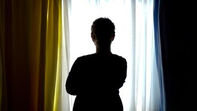 Rückansicht einer einsam wirkenden Frau, die an einem Fenster steht und hinausschaut.